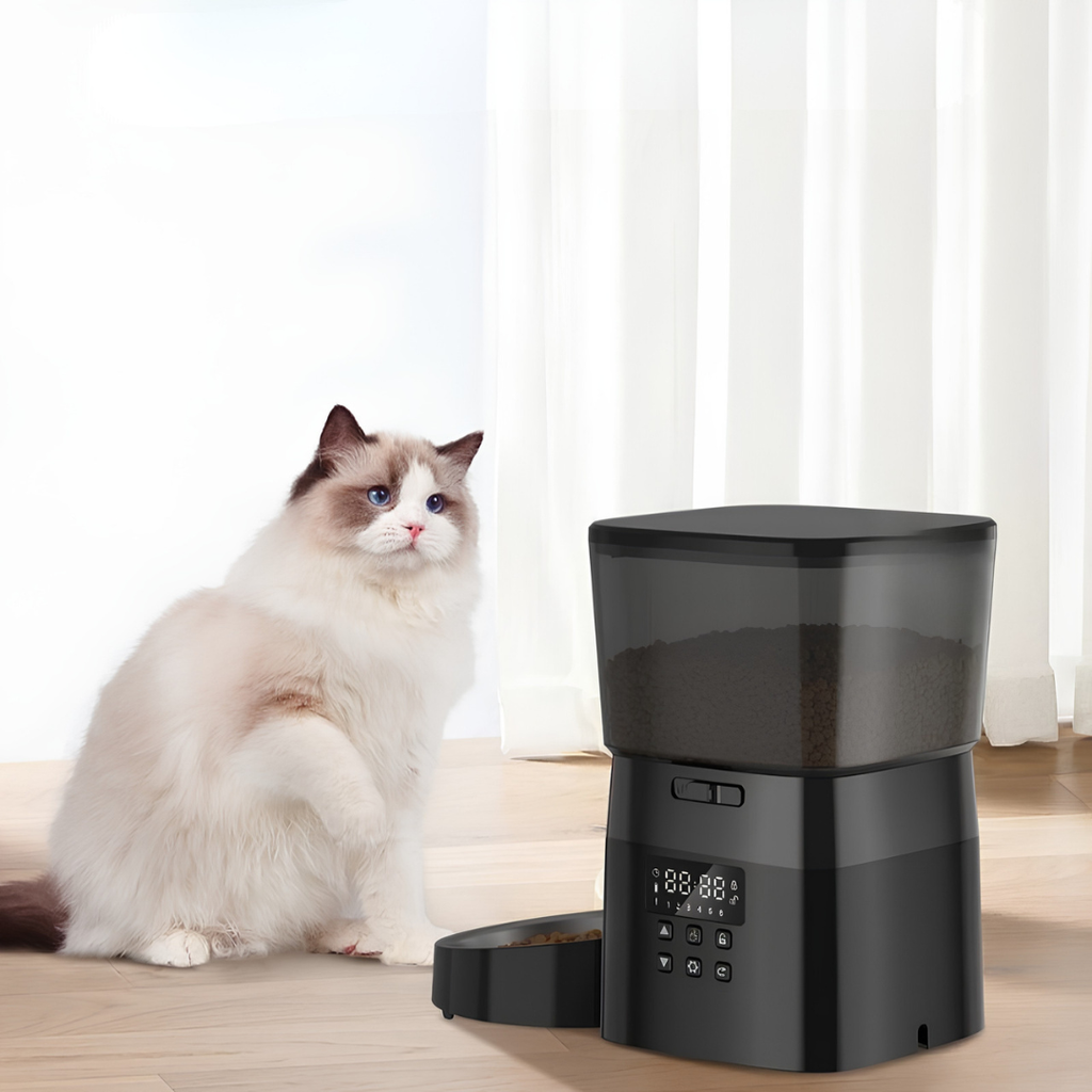 Cat sitting next to a black pet food dispenser on a wooden floor with white curtains in the background.